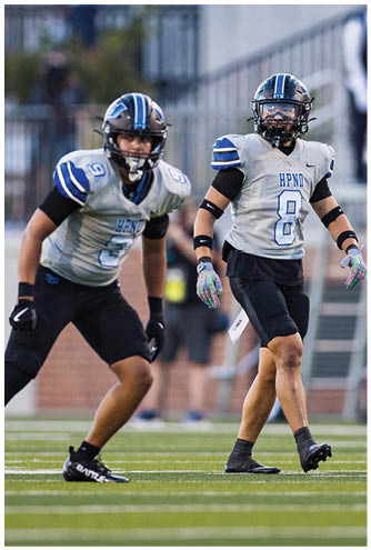 Two adults wearing football uniforms with helmets and gloves are standing on a football field during a game or practice. Description generated by AI
