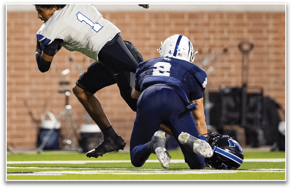 Two people in football uniforms are engaged in a play on a field, with one person falling forward and the other reaching toward a helmet on the ground. Description generated by AI