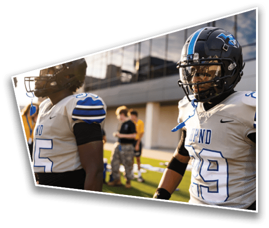 Two people in football uniforms and helmets stand on a field with blurred figures and a building in the background. Description generated by AI