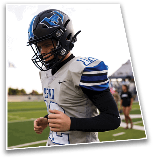 A person wearing a football helmet and gray jersey with blue and black stripes is standing on a football field with blurred spectators in the background. Description generated by AI