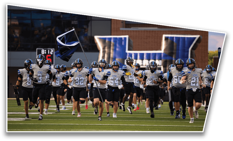 A group of people wearing football uniforms and helmets are running onto a field with one person holding a flag, and an inflatable tunnel is visible in the background. Description generated by AI