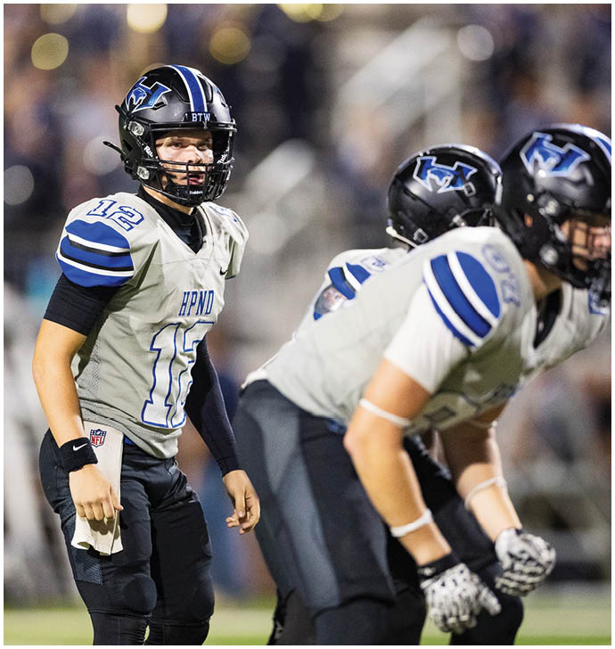 A person wearing a football uniform and helmet stands on a field next to two other players who are bent over in a ready position. Description generated by AI