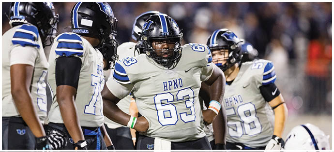 A group of people wearing football uniforms with helmets are gathered on a field, with one person in the center standing with hands on hips. Description generated by AI