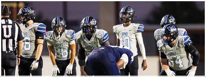A group of people wearing football helmets and uniforms are gathered on a field, appearing to prepare for a play during a game. Description generated by AI