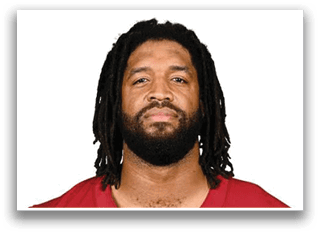 Person with shoulder-length dreadlocks and a beard wearing a red sports jersey looking directly at the camera against a white background. Description generated by AI