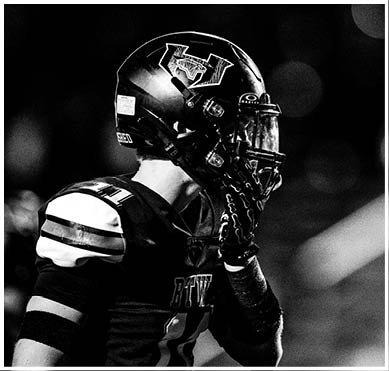 A close-up black and white photo of a football helmet with a logo on the side against a dark background with blurred light spots. Description generated by AI