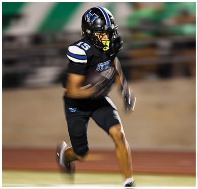 A person in a black football uniform with blue and white accents is running on a field during a game. Description generated by AI