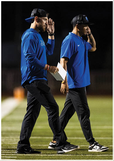 Two adults wearing blue shirts, black pants, and black caps walk side by side on a sports field at night, each holding a hand near their head. Description generated by AI