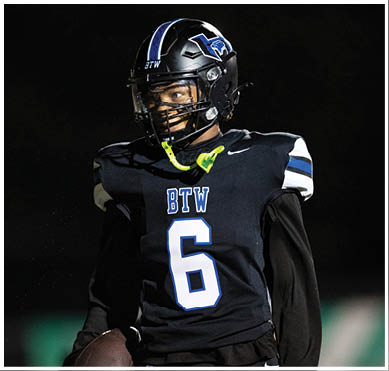 A person wearing a black football uniform with the number 6 and the letters BTW on the chest is holding a football and looking to the side on a dark field. Description generated by AI