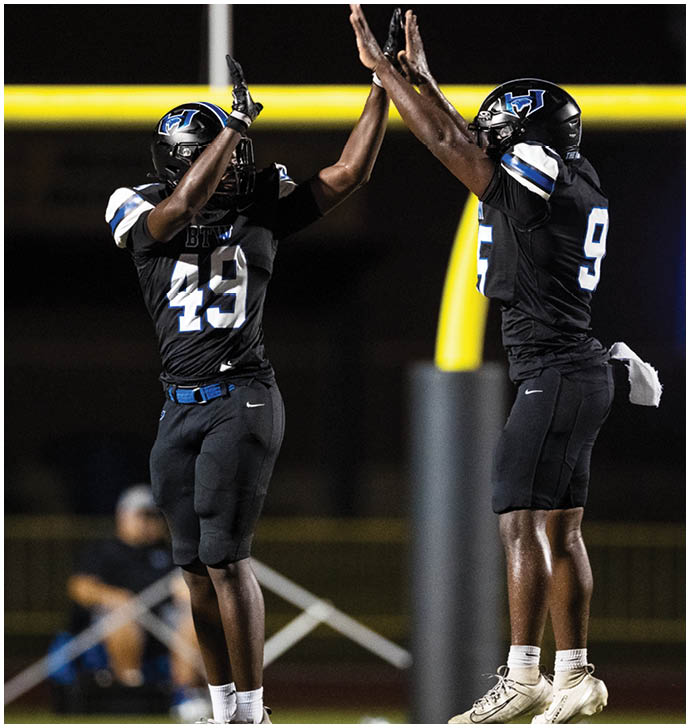Two people in black football uniforms with blue and white accents are giving each other a high five on a field with a yellow goalpost in the background. Description generated by AI