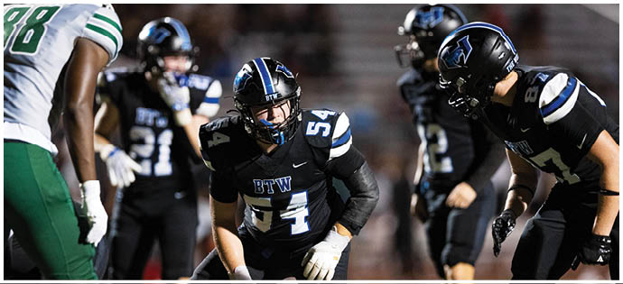 Four people wearing black football uniforms with blue and white stripes are on a field, appearing to prepare for a play. Description generated by AI