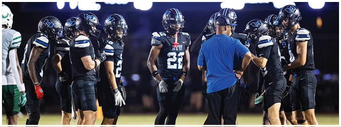 A group of people wearing black football uniforms and helmets are gathered on a field with one person in a blue shirt and headset appearing to give instructions. Description generated by AI