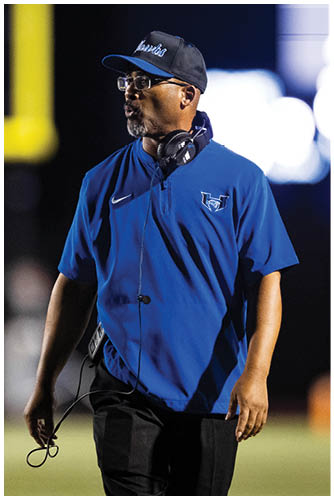 A person wearing a blue sports shirt, black pants, a cap, glasses, and headphones around their neck is standing on a field, appearing focused. Description generated by AI