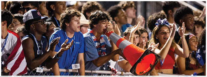 A group of people wearing blue and patriotic-themed clothing enthusiastically cheer and shout through a large orange megaphone at an outdoor event. Description generated by AI