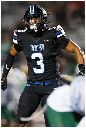 A person wearing a black football uniform with the number 3 and the letters BTW is running on a field with blurred players in the background. Description generated by AI