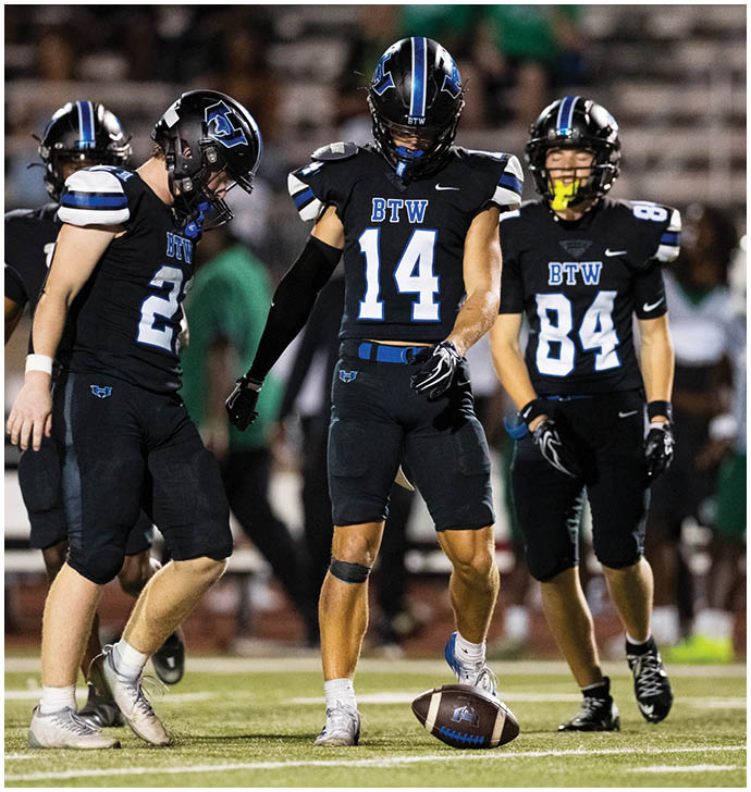 Three people in black football uniforms with blue and white accents stand on a field with one person preparing to kick a football. Description generated by AI