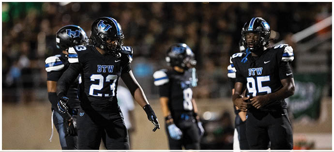 Four people in black football uniforms with blue and white accents stand on a field during a nighttime game. Description generated by AI