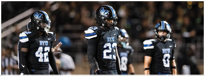 Four people wearing black football uniforms with blue and white accents stand on a field during a game. Description generated by AI