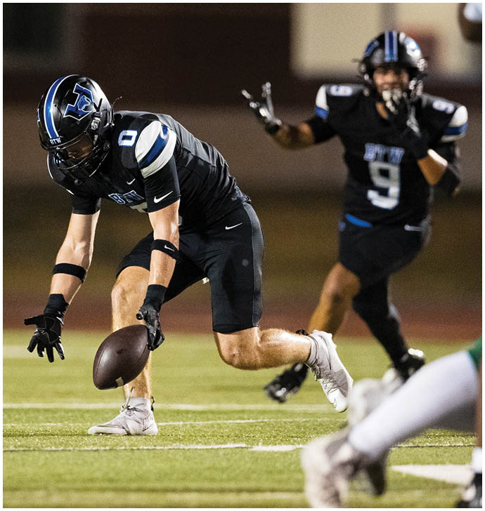Two people in black football uniforms are on a field with one bending down to catch a football while the other gestures behind. Description generated by AI