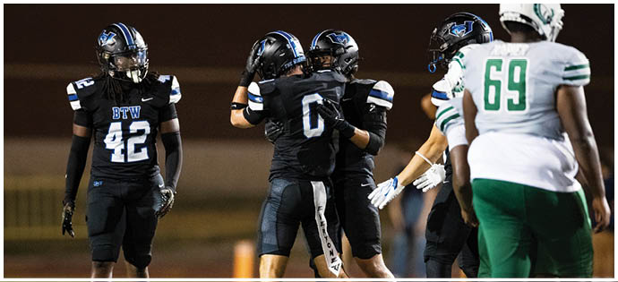Four football players in black uniforms and two players in white and green uniforms are on a field during a game, with two players in black celebrating by hugging. Description generated by AI