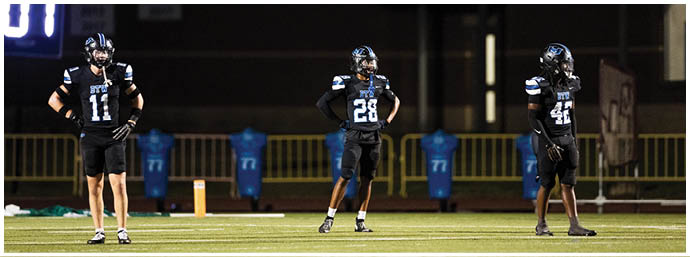 Three people wearing black football uniforms with helmets stand spaced apart on a field under stadium lights. Description generated by AI
