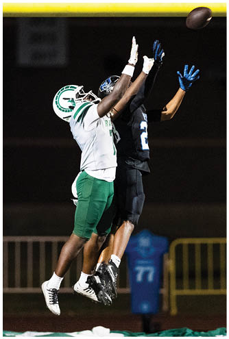 Two people in football uniforms jump with arms extended toward a football near the goalpost on a field at night. Description generated by AI