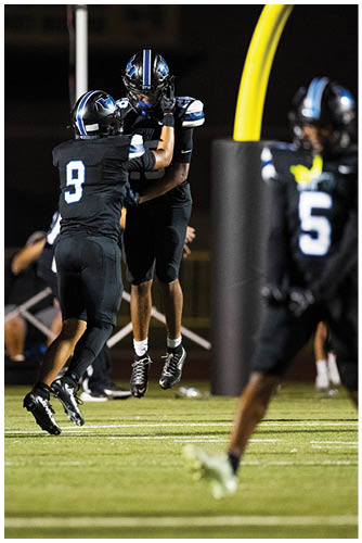 Two people in black football uniforms with blue and white helmets celebrate by jumping and touching hands on a football field at night. Description generated by AI