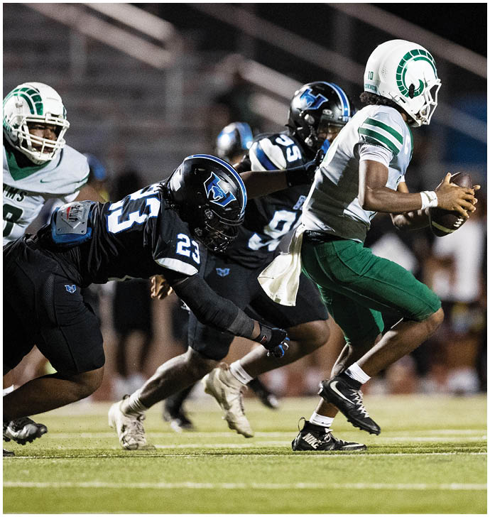 A person in a white and green football uniform is holding a football while two people in black and blue uniforms are attempting to tackle them on a football field. Description generated by AI