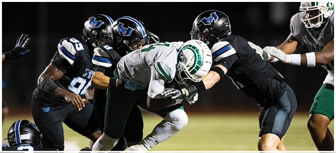 A group of people in football uniforms are engaged in a tackle during a nighttime game on a field. Description generated by AI