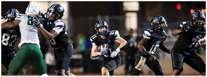 People in football uniforms are actively playing on a field with one person running while holding the ball and others blocking or preparing to move. Description generated by AI