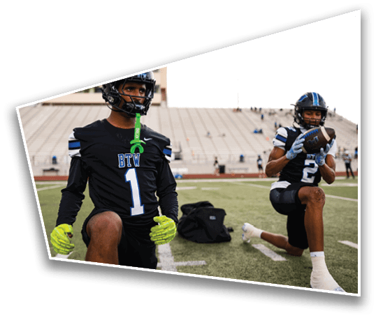 Two people in football uniforms are on a field, with one kneeling and holding a football while the other stands nearby. Description generated by AI