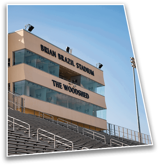 A sports stadium with empty bleachers and a beige press box labeled Brian Brazil Stadium and The Woodshed under a clear blue sky. Description generated by AI