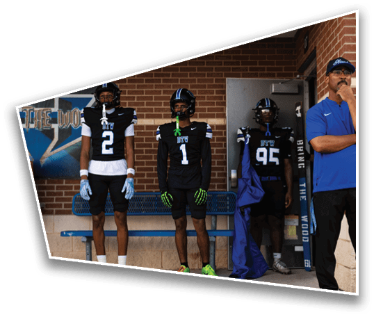 Three people in black football uniforms stand next to a person in a blue shirt and cap against a brick wall near a bench and a door. Description generated by AI