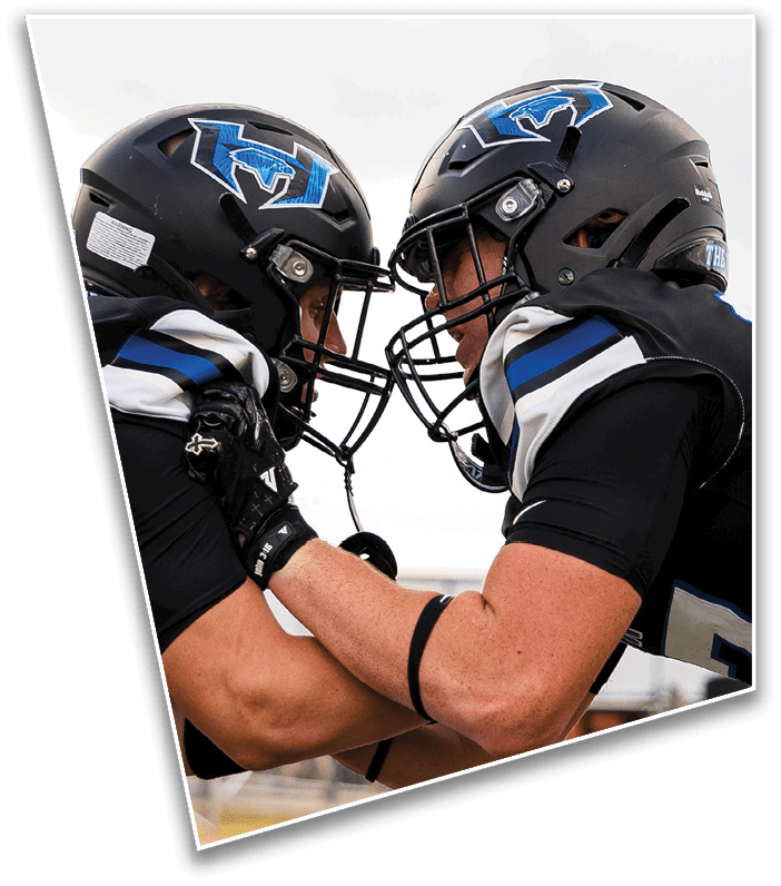 Two football players in black helmets and uniforms face each other closely with their helmets touching, appearing to engage in a competitive or intense moment. Description generated by AI