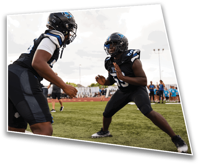 Two people in football gear are positioned on a field facing each other with a group of people in blue shirts standing in the background. Description generated by AI