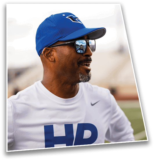 A person wearing a blue cap, sunglasses, and a white shirt with “HD" printed on it is smiling and looking to the side on a sports field. Description generated by AI