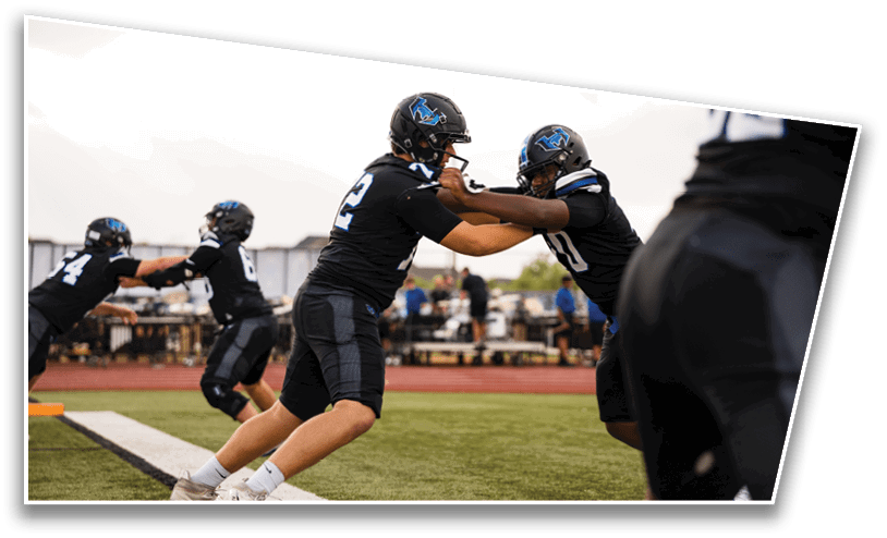 People in black football uniforms and helmets are practicing blocking drills on a field near the sideline. Description generated by AI