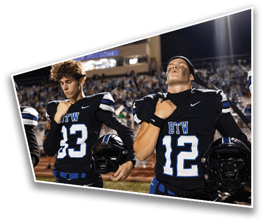 Two people in black football uniforms with blue and white accents stand on a field holding helmets and placing their hands over their chests during a nighttime event. Description generated by AI
