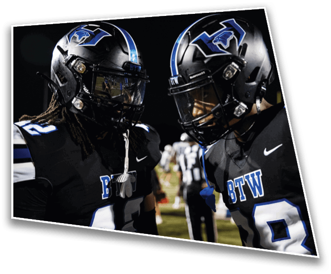 Two people wearing black football uniforms and helmets with a blue and white logo are facing each other on a field at night. Description generated by AI