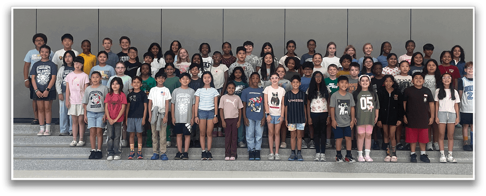 A large group of children and adults stand closely together indoors against a plain wall, many smiling and wearing casual clothing. Description generated by AI