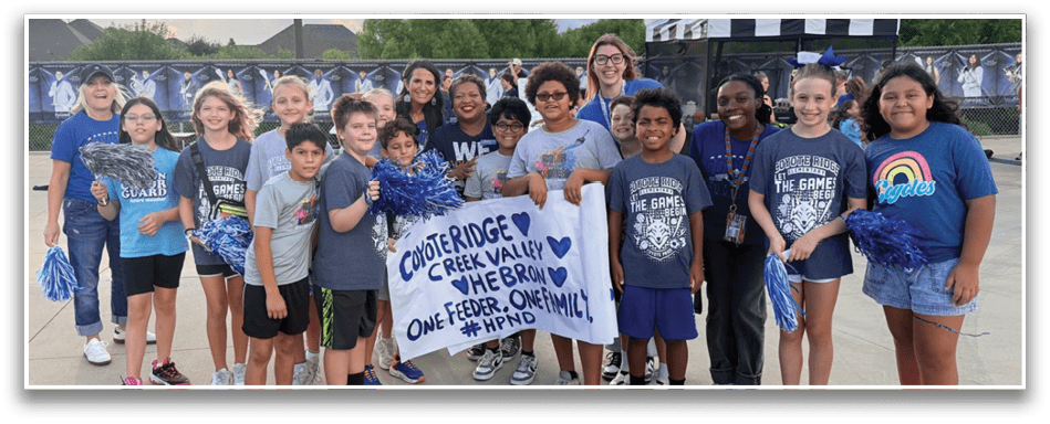 A group of children and adults stand outdoors holding a sign that reads “Coyote Ridge Creek Valley We Bear One Feather, One Family #HPND" with some wearing school-themed shirts and holding blue pom-poms. Description generated by AI
