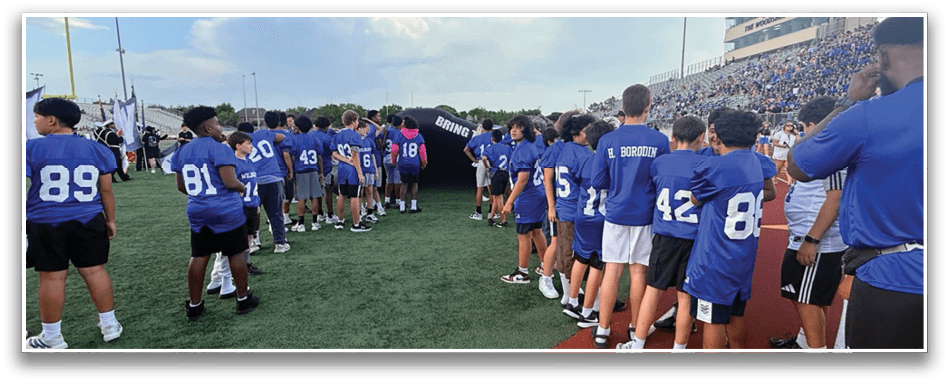 People wearing blue sports jerseys are lined up on a grassy field near a stadium filled with spectators under a partly cloudy sky. Description generated by AI