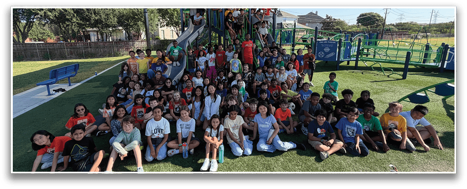 A large group of children sitting closely together on grass outdoors, smiling and posing for a group photo on a sunny day. Description generated by AI