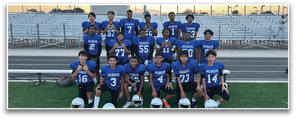 A group of people wearing blue football jerseys with “WILDCATS" printed on them are posing together on a sports field with helmets placed in front of them and empty bleachers in the background. Description generated by AI