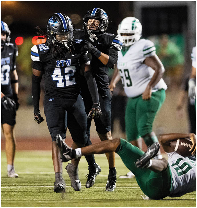 Two people in black football uniforms are standing while one person in a green and white football uniform is falling backward on the field with two other players in green and white standing in the background. Description generated by AI
