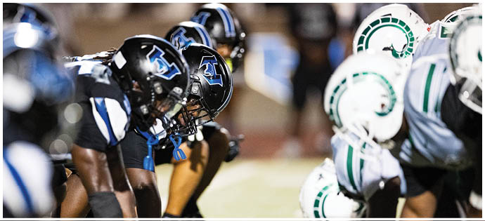 Two opposing American football teams in helmets and uniforms are lined up facing each other on the field, ready for the next play. Description generated by AI