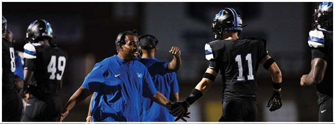 A person in a blue shirt is giving a high-five to a person in a black sports uniform with the number 11 on the back, while other people in black uniforms stand nearby on a sports field. Description generated by AI