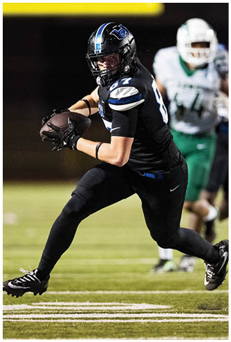 A person in a black sports uniform is running on a football field with two other people in green and white uniforms visible in the background. Description generated by AI