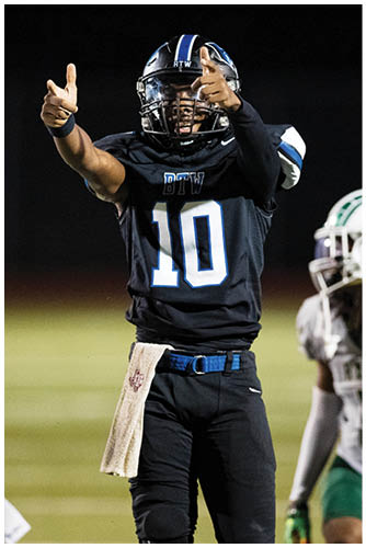 A person wearing a black football uniform with the number 10 is signaling with both hands while two other people in white and green football uniforms stand nearby on a field. Description generated by AI