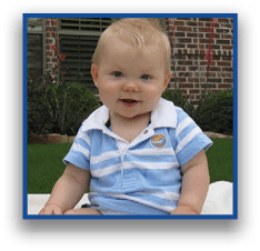 A child wearing a blue and white striped romper with a collar is sitting on a white blanket outdoors on grass with plants and a brick wall in the background. Description generated by AI